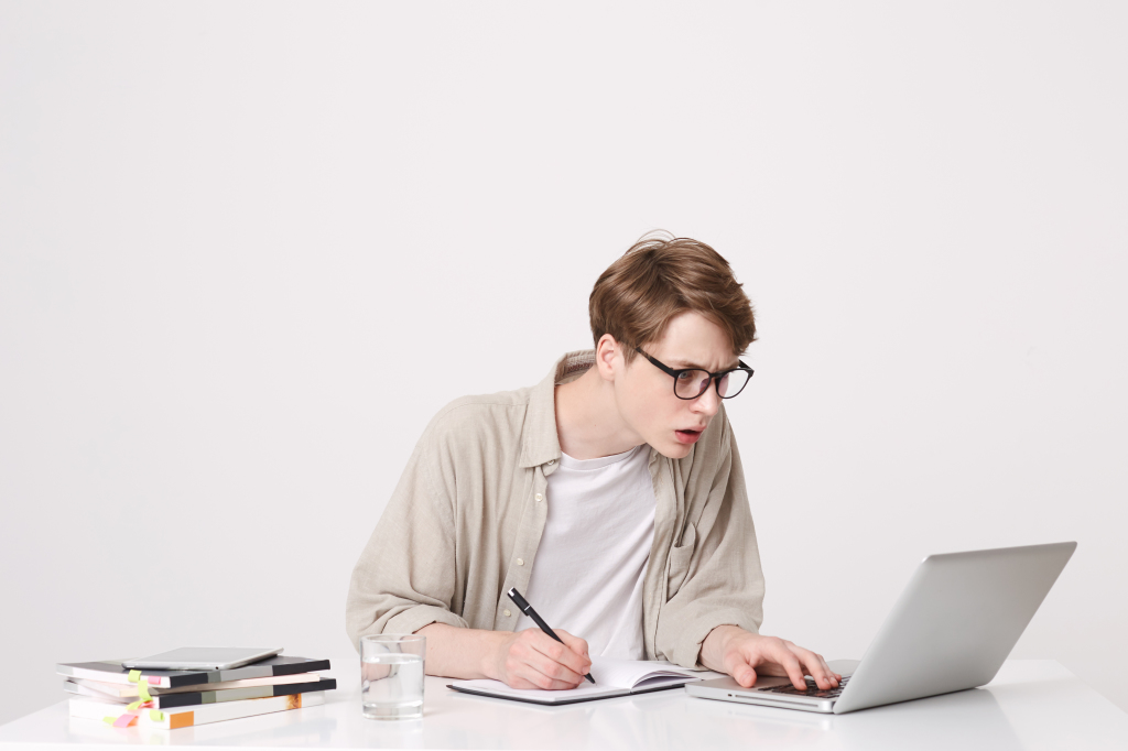 Closeup of concentrated young man student wears glasses and beige shirt sitting and working with laptop computer and notebooks at the table isolated over white background